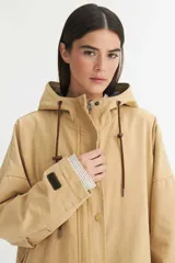 Parka color beige de canvas con forro de nylon, cierre frontal con cremallera y broches a presión, capucha con cordón ajustable y puños con detalle de rayas.