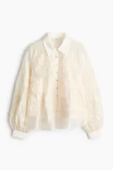 Blusa oversized de tejido transparente color crudo, con detalles bordados en relieve, cuello camisero, cierre frontal de botones y mangas largas abullonadas con puños ajustados.