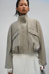 Chaqueta corta de ante con cuello alto, manga larga y cierre frontal con cremallera y botones a presión. Presenta bolsillos frontales con solapa y bajo abullonado con pliegues.