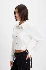 Camisa corta estilo corset de color blanco, con cuello clásico, cierre frontal de botones y detalles de cordones laterales ajustables. Presenta pinzas frontales y traseras para un ajuste entallado.