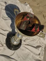 Sandalias de cuero con tiras anchas en color negro y beige, con un detalle circular metálico en el centro. La suela tiene un borde trenzado en blanco y negro.