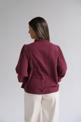 Blusa color bordó con bordados florales en tono crema en la parte superior frontal. Presenta cuello redondo con pequeña abertura, cierre frontal con botones y mangas largas acampanadas con puños ajustados.