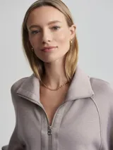 Campera deportiva con cierre frontal, cuello alto tipo chimenea y mangas raglán. Es de corte holgado y presenta bolsillos de parche delanteros. El color es un tono neutro entre gris y rosado (taupe).