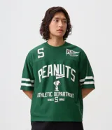 Remera de corte boxy y estilo deportivo, color verde oscuro, con diseño inspirado en el fútbol americano de Peanuts. Presenta cuello redondo, mangas cortas con franjas blancas horizontales y estampas localizadas de Snoopy en el frente.
