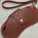 Estuche para lentes de diseño ergonómico y minimalista, confeccionado en cuero sintético con textura granulada. Cuenta con cierre magnético frontal, costuras a la vista en color contrastante y una correa de mano extraíble.