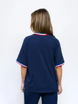 Remera blanca de manga corta con cuello redondo y puños ribeteados en rojo, blanco y azul. Presenta el escudo del Club Nacional de Football bordado en el pecho izquierdo y el logo de Umbro bordado en el pecho derecho.