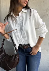 Camisa blanca de corte clásico con detalles de encaje floral vertical en la parte frontal y en los puños. Presenta cuello camisero, cierre frontal con botones y mangas largas con terminación elástica.