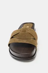 Sandalia plana con tiras cruzadas de gamuza color verde oliva, con detalles de tachas metálicas doradas en los laterales y suela de madera oscura.