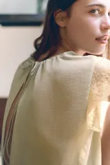 Body tipo blusa de georgette color verde oliva, con cuello redondo ajustable mediante lazada, mangas muy cortas con detalle de encaje y pliegues verticales en el delantero. Presenta una base de encaje en la parte inferior y cierre con botones a presión en el bajo.
