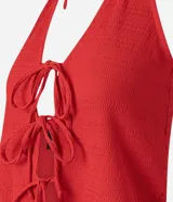 Vestido midi rojo, con textura plisada o arrugada, escote halter en V y detalle de amarras ajustables en el centro del busto que generan una abertura frontal. La falda presenta un fruncido lateral y una abertura alta.