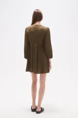 Vestido corto de color verde militar, con escote en V profundo, mangas largas con puños elásticos y detalles de encaje y bordados en el frente. Presenta una falda con vuelo y textura de pequeños puntos en relieve.