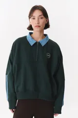 Buzo estilo rugby de corte oversized, color verde oscuro, con cuello tipo camisa en denim azul claro y cierre frontal parcial. Presenta un recorte horizontal en el pecho, logo bordado y tiras laterales en las mangas en contraste.