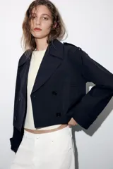 Trench corta de color negro con cuello de solapa, cierre frontal cruzado con botones y mangas largas con trabillas ajustables. Cuenta con bolsillos delanteros de vivo.