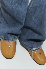 Zapato tipo bailarina plana de piel en color tostado, con pespuntes en el cuerpo, detalle metálico en la parte delantera y acabado en punta redonda.