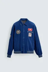 Cazadora estilo varsity confeccionada en pana de algodón azul, con interior ligeramente acolchado. Presenta cuello solapa, cierre frontal con botones a presión, bolsillos laterales de vivo y acabados en rib a rayas en puños y cintura. Incluye múltiples parches bordados decorativos en el frente.