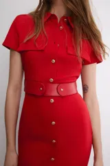 Vestido midi de color rojo, con cuello solapa, manga corta y cierre frontal de botones metálicos. Presenta bolsillos decorativos en el pecho y un cinturón a tono en la cintura.