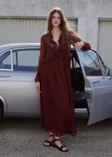 Vestido largo de tejido fluido color bordó, de diseño evasé con escote en pico y mangas largas con puños elásticos. Presenta detalles de volantes en el cuello y forro interior.