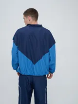 Campera deportiva Umbro modelo Beat, confeccionada en nylon ligero con diseño de bloques de color en blanco, azul marino y verde azulado. Presenta cierre frontal, cuello alto y elásticos en puños y cintura.