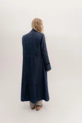 Tapado largo de jean azul oscuro, con cuello solapa y puños con detalle de cinta de terciopelo negro. Presenta costuras visibles que definen paneles y un logo bordado en la espalda.