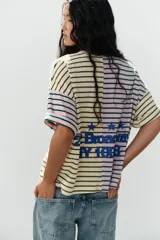Camiseta oversize con diseño bicolor a rayas horizontales, combinando un lado en tonos rosado y verde oscuro, y el otro en beige y marrón. Cuenta con cuello redondo, mangas cortas amplias y texto bordado en azul en el pecho.
