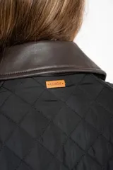 Campera negra de diseño capitoneado con cuello de solapa en contraste color marrón. Presenta cierre frontal con cremallera y botones a presión, mangas largas con puños ajustables y un corte recto.