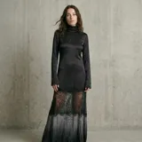 Vestido largo de satén negro con cuello alto y mangas largas. Presenta un diseño con una franja de encaje negro transparente en la parte inferior, que conecta el cuerpo de satén con una falda de satén en degradado gris oscuro.