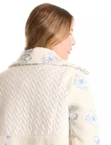 Chaqueta de corte relajado confeccionada en tejido tipo corderito color crema, combinada con paneles de matelassé en el frente y bolsillos. Presenta un delicado bordado floral en tonos azules sobre el corderito, cierre frontal metálico dorado y bolsillos de parche delanteros.