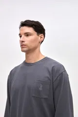 Remera de manga larga en color gris oscuro, confeccionada en algodón pesado con textura suave. Presenta un bolsillo frontal en el pecho con un pequeño bordado de la marca y puños elásticos en las mangas.