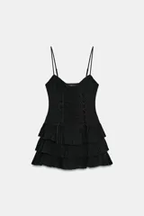 Vestido corto negro de tirantes finos ajustables y escote en V. Presenta un diseño ajustado con drapeado central y cordones cruzados laterales tipo corsé, y falda corta con tres capas de volantes escalonados.