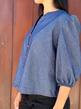 Camisa de jean azul con cuello mao, cierre frontal con botones rojos y mangas tres cuartos abullonadas con puños elásticos.