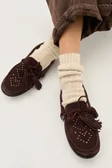 Mocasín de cuero color marrón oscuro, con diseño tipo mocasín de gamuza, detalle de flecos con borlas en el empeine y tachas metálicas pequeñas distribuidas en la parte frontal.