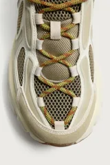 Zapatilla deportiva tipo running con diseño de suela voluminosa e irregular. Presenta una combinación de piezas en tonos beige y crema, con paneles de malla y cordones jaspeados.