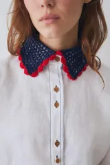 Camisa de lino blanca con cuello y puños tejidos a mano en crochet azul con borde rojo. Presenta cierre frontal con botones de madera, bolsillo frontal y costuras en contraste.