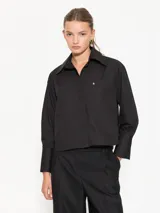 Camisa negra de poplin, corte recto, con cuello clásico, cierre frontal con botones ocultos, dos bolsillos de parche en el pecho, mangas largas con puños abotonados y ruedo redondeado.