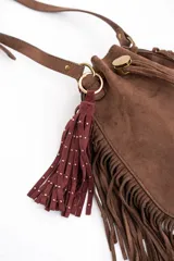 Cartera tipo bucket color marrón, confeccionada en material sintético con acabado gamuzado. Presenta flecos largos en los laterales y la base, cierre mediante cordón ajustable y correa larga regulable para llevar al hombro o cruzada.