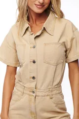 Mono corto color beige, estilo denim, con cuello camisero, cierre frontal con botones, mangas cortas y bolsillos en el pecho y laterales.