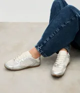 Championes de estilo urbano inspirados en botines de fútbol, confeccionados en poliuretano plateado con paneles de malla transpirable. Presentan cierre con cordones, punta redondeada y suela plana de goma en color marrón.
