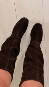 Bota de caña alta de gamuza en color marrón chocolate, con taco cuadrado y puntera cuadrada. Cuenta con un diseño holgado en la caña y detalle de trabilla decorativa en el tobillo.