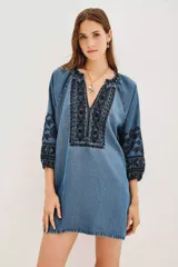 Vestido corto de tela tipo denim con bordados geométricos en color negro en el pecho y las mangas. Presenta escote en V, mangas tres cuartos con puños elásticos y un corte holgado.