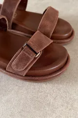 Sandalia plana color marrón de cuero vacuno, con tres tiras ajustables con hebillas y cierre de velcro, y base acolchada.