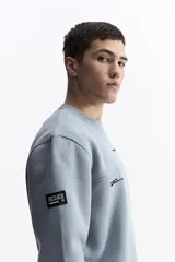 Buzo de algodón color celeste, con cuello redondo, puños y bolsillos laterales. Presenta estampados con texto en el pecho y un parche en la manga.