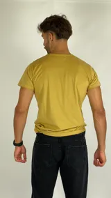 Remera de manga corta color amarillo mostaza con cuello redondo. Presenta un estampado frontal con el texto 'Risk Or Lost' y un gráfico de un ave con sombrero sobre un sol poniente.