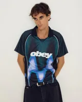 Camiseta de fútbol Obey con diseño estampado de llamas en tonos rojos, naranjas y amarillos. Presenta cuello polo negro, escote en V y paneles laterales en color negro.