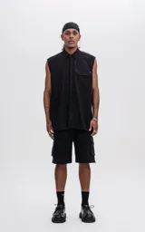 Camisa sin mangas de corte oversize, color negro, confeccionada en tejido twill. Presenta cuello clásico, cierre frontal oculto y un bolsillo tipo cargo en el pecho con detalle de remaches metálicos.