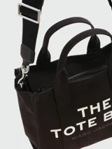 Bolso tipo tote de lona color negro con estampado frontal en blanco que dice 'THE TOTE BAG' y 'MARC JACOBS'. Cuenta con doble asa superior, correa ajustable y extraíble para llevar al hombro y cierre superior con cremallera.