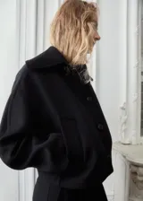 Chaqueta negra de corte corto y holgado, confeccionada con mezcla de lana. Cuenta con cuello ancho y solapa de piel sintética en tono beige claro, cierre frontal con botones oscuros y mangas largas amplias.