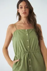 Monoprenda corta de color verde oliva, con tirantes finos ajustables, cierre frontal con botones y cintura elástica con cordón ajustable. Cuenta con bolsillos laterales.