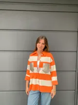 Camisa tejida a rayas horizontales color naranja y blanco, con cuello camisero, mangas tres cuartos y bolsillos frontales con solapa en color dorado.