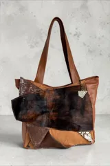 Cartera tipo tote de cuero en tono marrón, combinada con apliques de pelo vacuno animal print, detalles metalizados y dije colgante triangular.