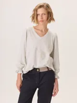 Remera de manga larga color gris claro melange, con escote en V ribeteado y un sutil bordado en el centro del pecho. Las mangas tienen un ligero volumen en el hombro y puños fruncidos con elástico.